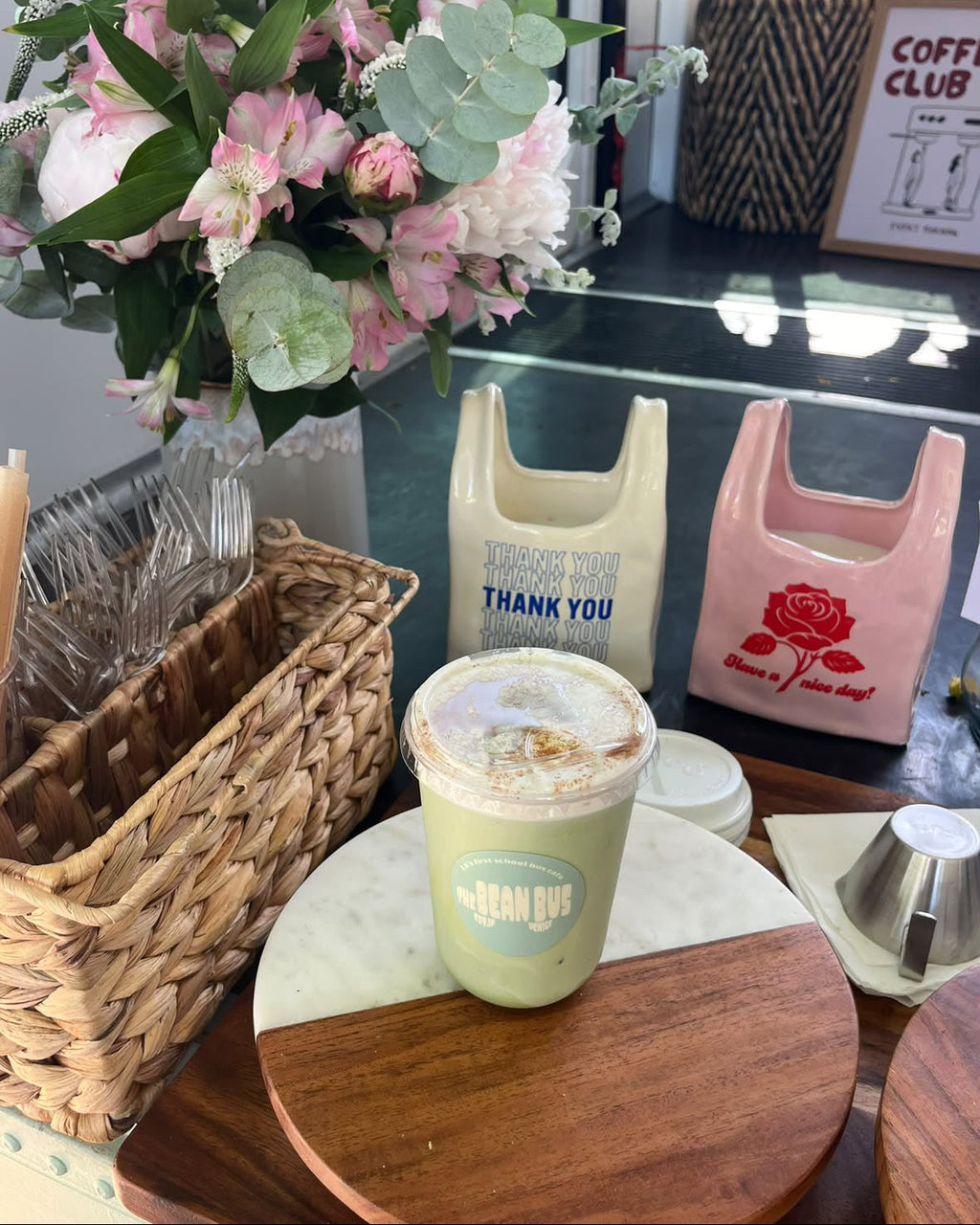 A green iced coffee drink in a clear plastic cup with a blue 'The Bean Bus' logo, displayed on a wooden and marble coaster. The setting is a cafe table decorated with a large pink and green floral arrangement, two reusable shopping bags, and a wicker basket.
