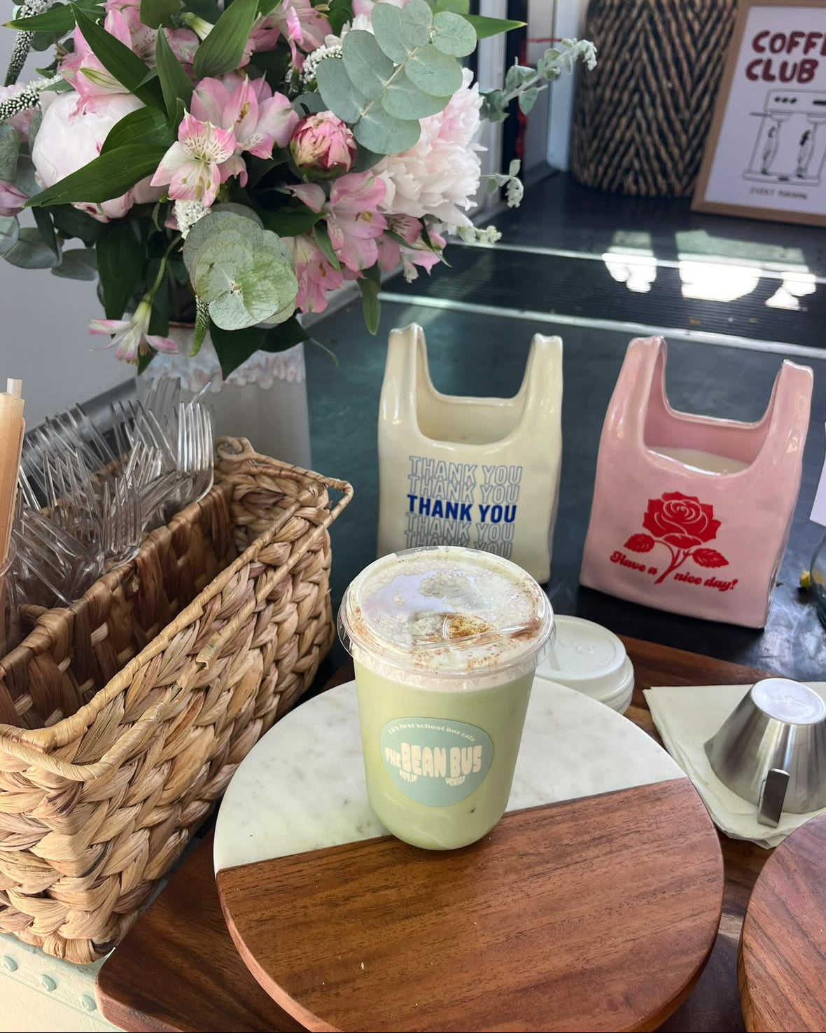 A green iced coffee drink in a clear plastic cup with a blue 'The Bean Bus' logo, displayed on a wooden and marble coaster. The setting is a cafe table decorated with a large pink and green floral arrangement, two reusable shopping bags, and a wicker basket.