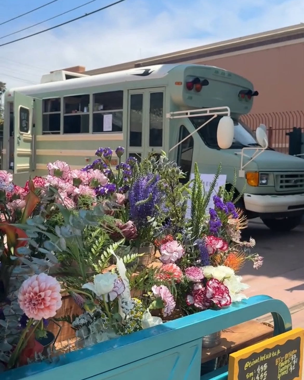 A turquoise blue pickup truck bed filled with a colorful assortment of fresh flowers, parked outdoors in front of a vintage light green bus under a blue sky.