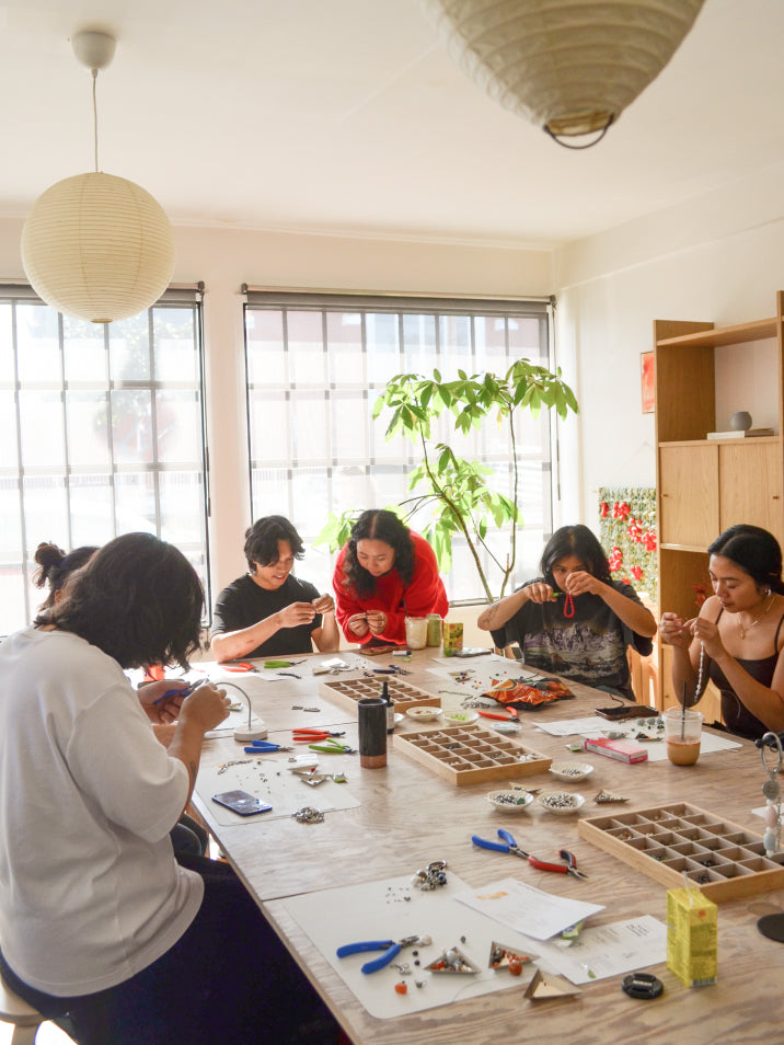 Group of people sitting around a table making jewelry in a room with plants and large windows.