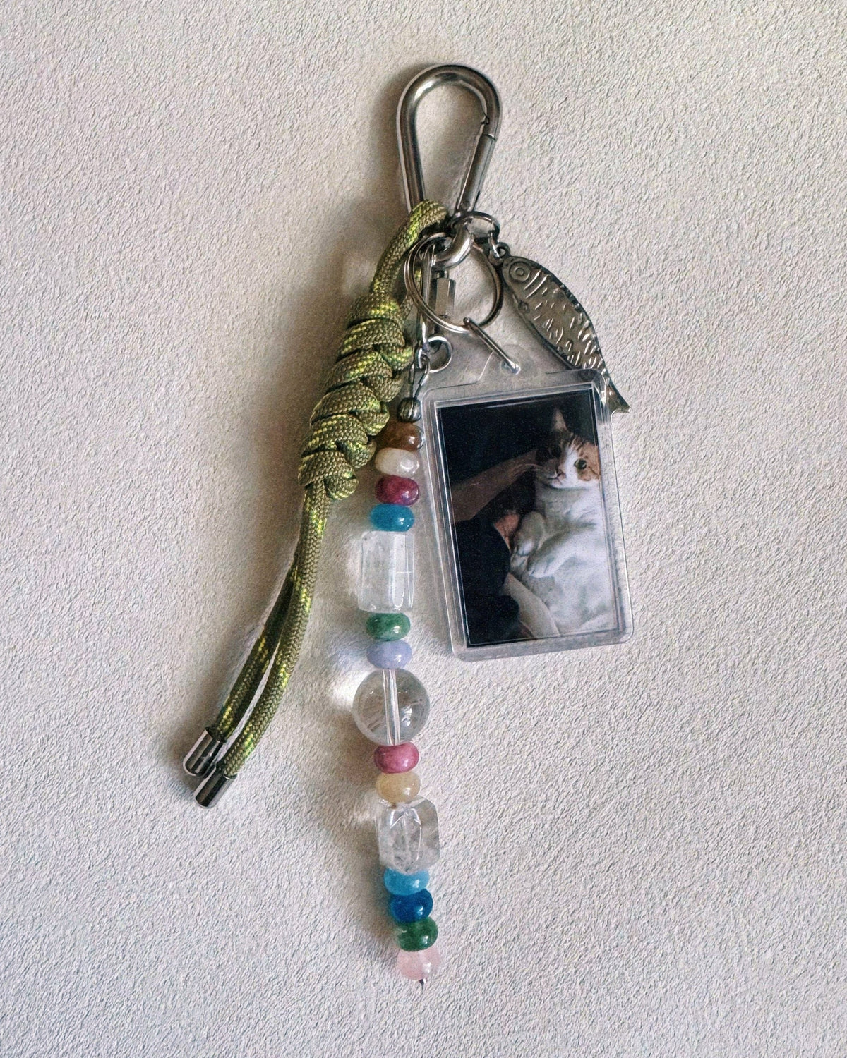 A handmade keychain featuring a silver carabiner, a knotted olive green cord, a silver fish charm, a clear acrylic photo holder with a cat picture, and a beaded drop with multicolored beads on a textured off-white background.
