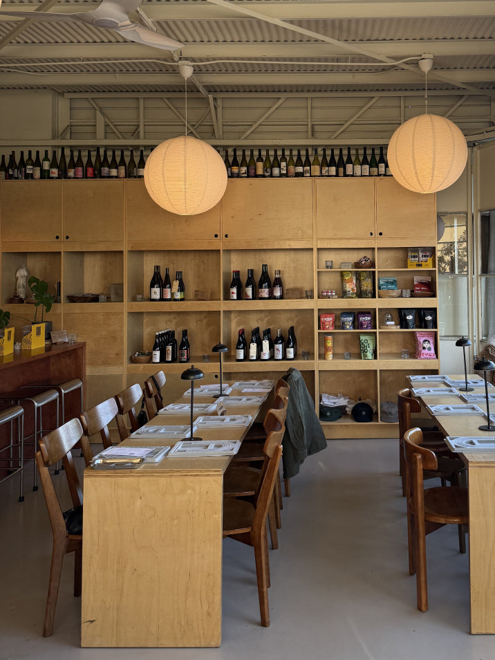 A large, bright meeting or workshop room with long wooden tables and chairs, built-in shelving filled with wine bottles, two large hanging paper lantern lights, and an industrial ceiling.