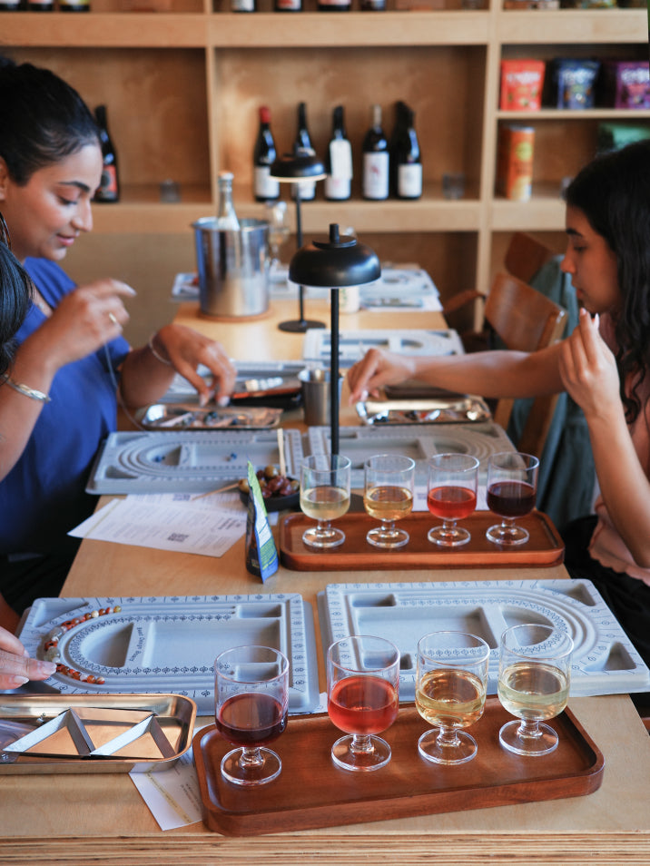 Two women seated at a wooden table in a wine tasting or restaurant setting, sampling different colored wines from small glass cups arranged on wooden trays. The scene includes shelves with wine bottles in the background.