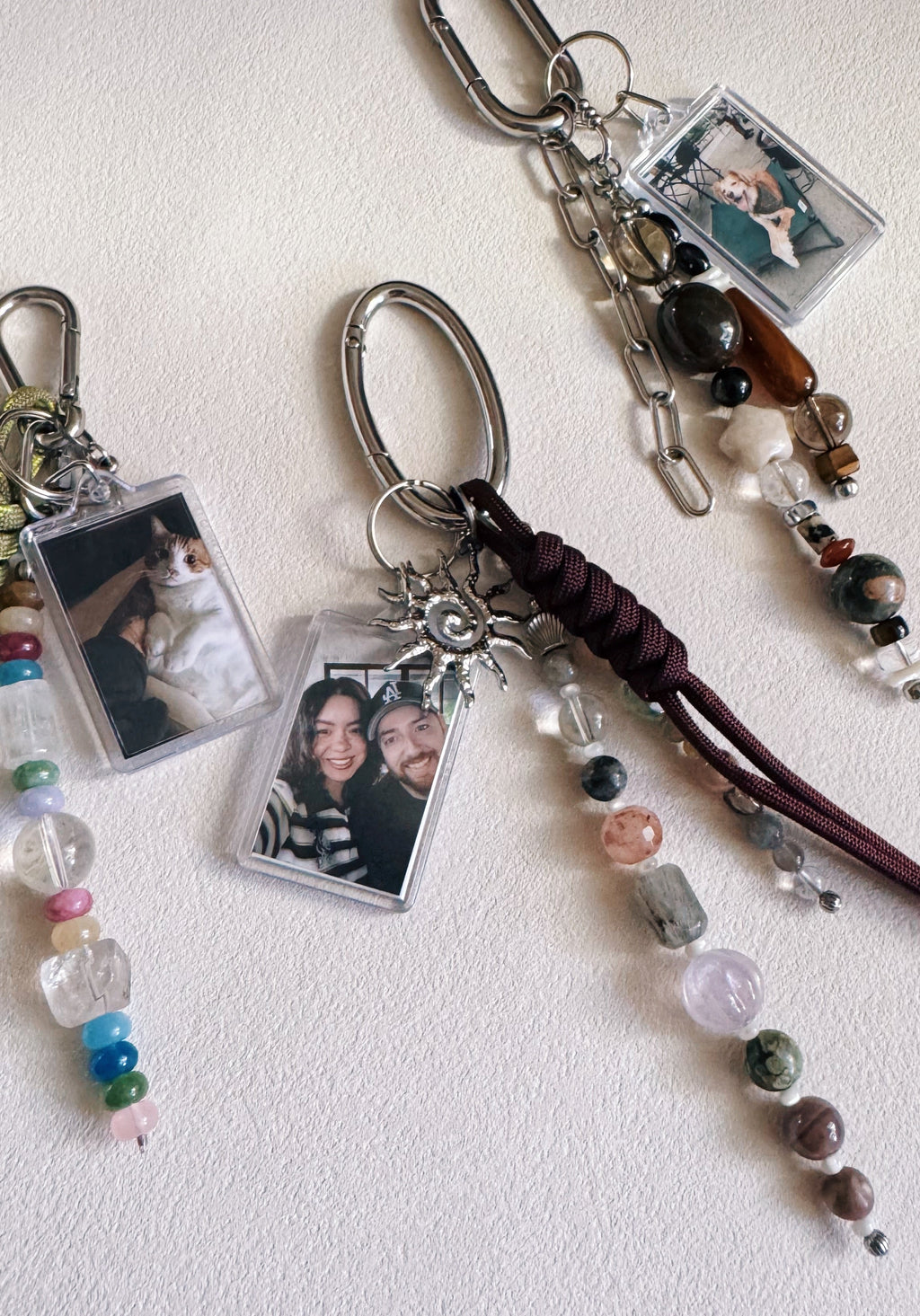 Three personalized keychains with clear photo frames, silver keyrings, colorful beads, and braided dark brown cords, arranged on a light textured background.