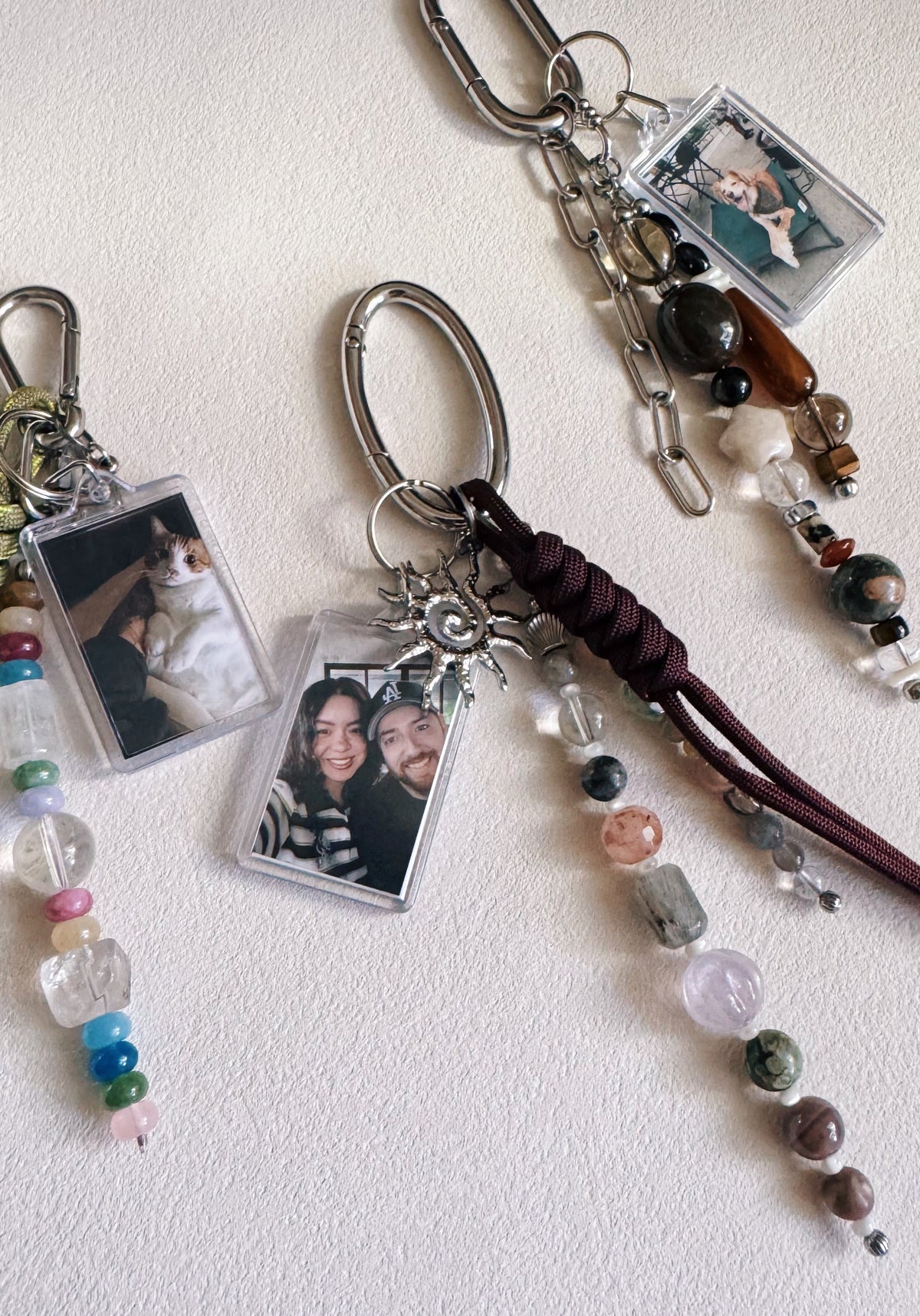 Three personalized keychains with clear photo frames, silver keyrings, colorful beads, and braided dark brown cords, arranged on a light textured background.