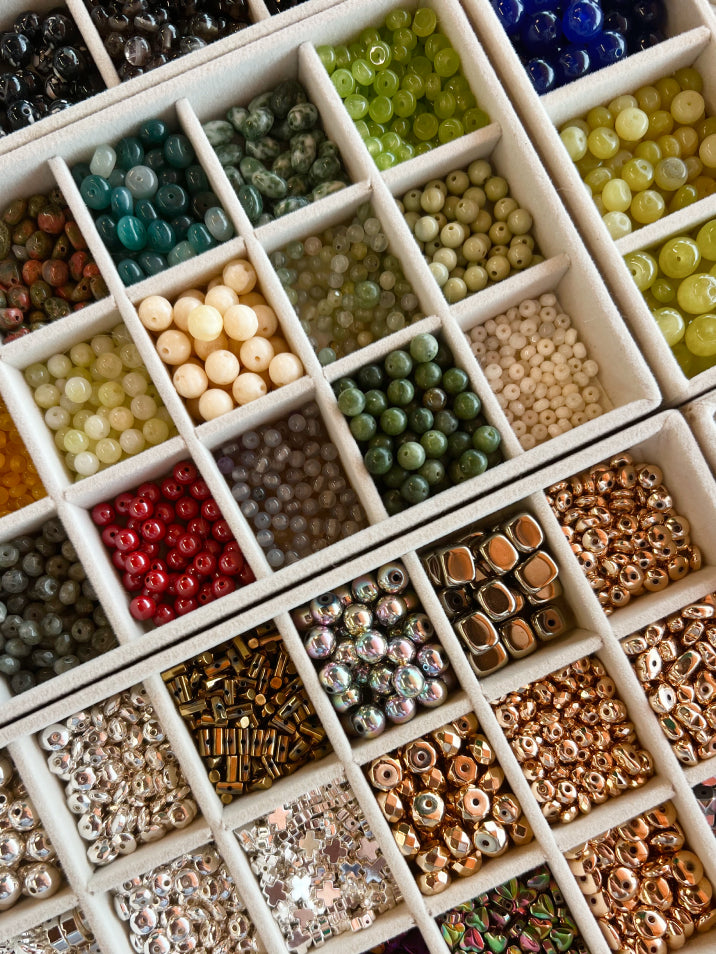 An overhead view of a white compartment organizer filled with assorted craft beads in many colors and shapes, including round, oval, and metallic styles.