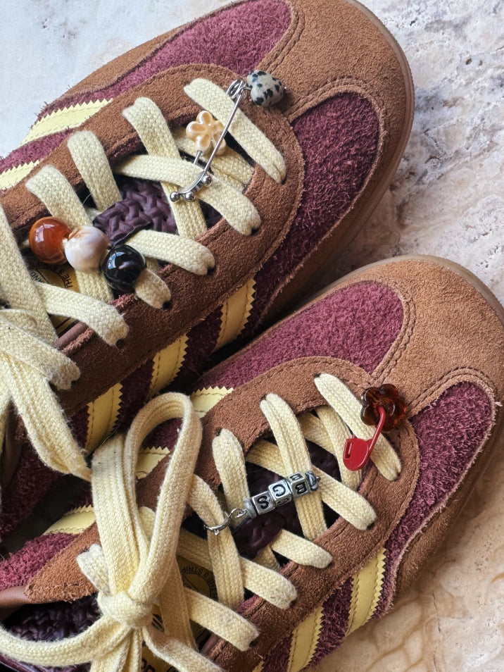 A close-up of a pair of brown suede sneakers with burgundy mesh panels, cream laces, and decorative bead and charm details on the laces, set on a light stone surface.